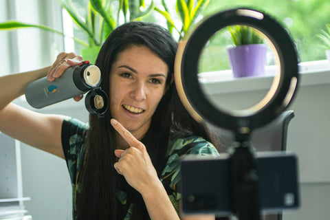 Gredzena formas lampa ar USB kabeli un telefonu turetaju, fonā cilvēks filmē video